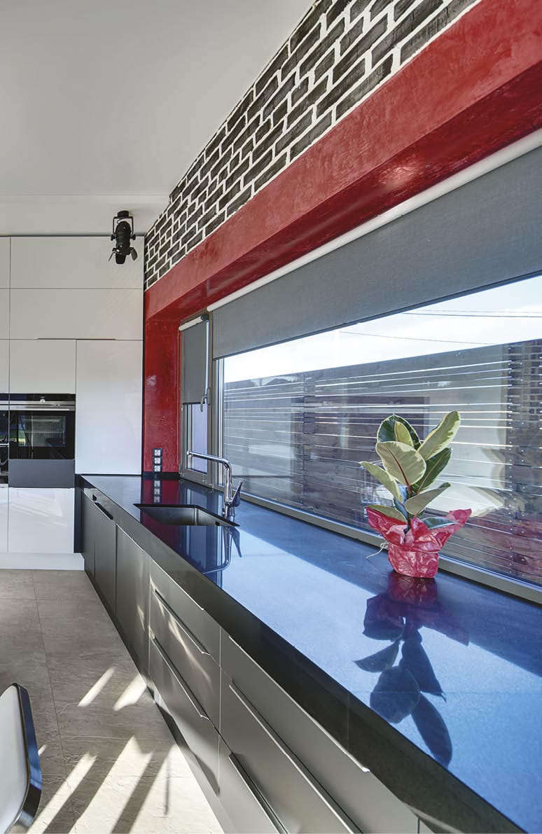 Kitchen zone in a modern style with a built-in oven, a kitchen island, a sink, a chair and a large windows with curtains. On the left there is a red-cream stair, plants in the pots, a door.